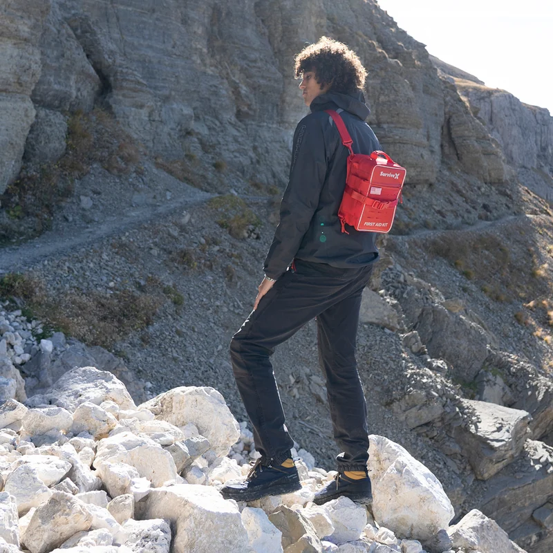 Hiker carrying a red SurviveX first aid kit in a mountain setting